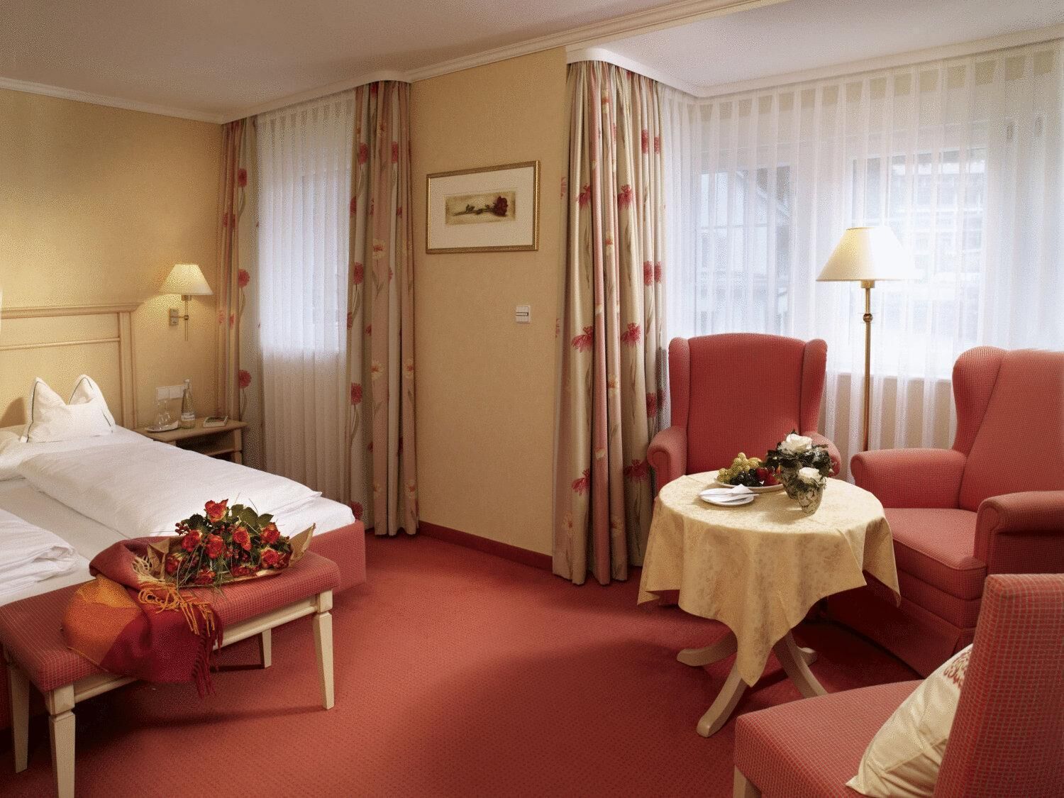 A hotel room with a bed, a bedside table with a lamp, a small table with a vase of flowers, and two armchairs.