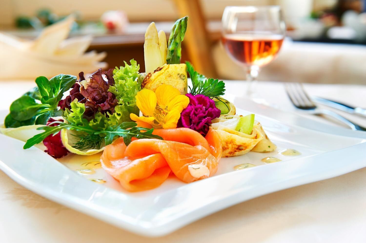 Een vierkant wit bord met een salade, zalm en een glas wijn.