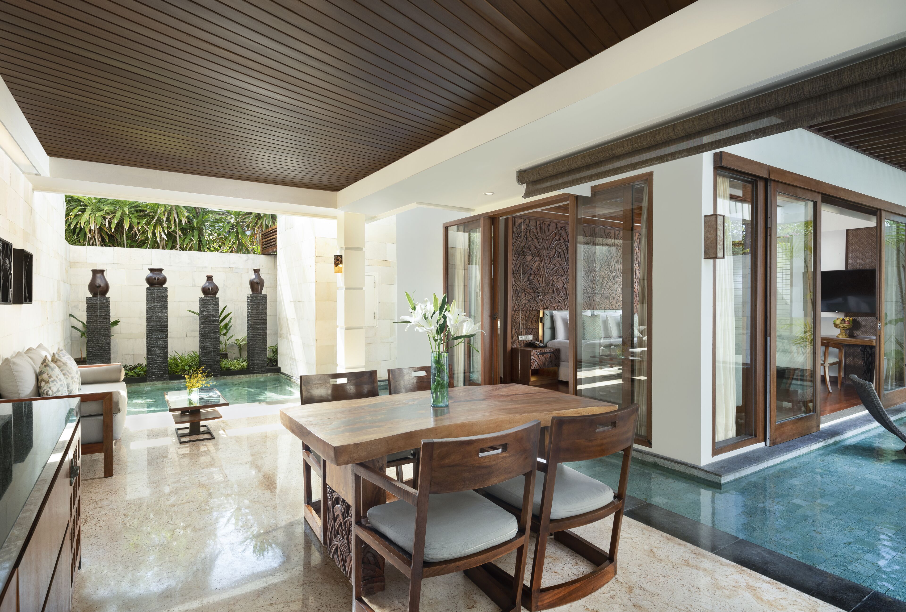 Um espaço amplo e interno com uma mesa de jantar de madeira, cadeiras e uma área de estar com sofá. Uma piscina é visível através das portas de vidro.