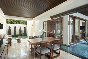 Um espaço amplo e interno com uma mesa de jantar de madeira, cadeiras e uma área de estar com sofá. Uma piscina é visível através das portas de vidro.