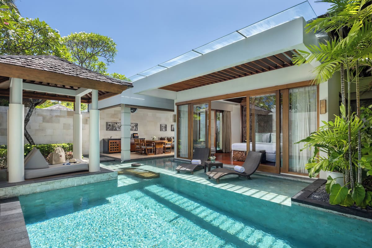 A modern pool area with lounge chairs, a covered seating area, and a glass-enclosed room with a bed.