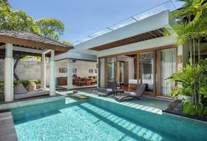 Un área de piscina moderna con sillones de descanso, una zona de estar cubierta y una habitación con paredes de vidrio y una cama.