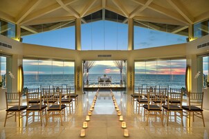Un amplio salón con suelo reflectante, filas de sillas de madera y grandes ventanales con vista al mar al atardecer.