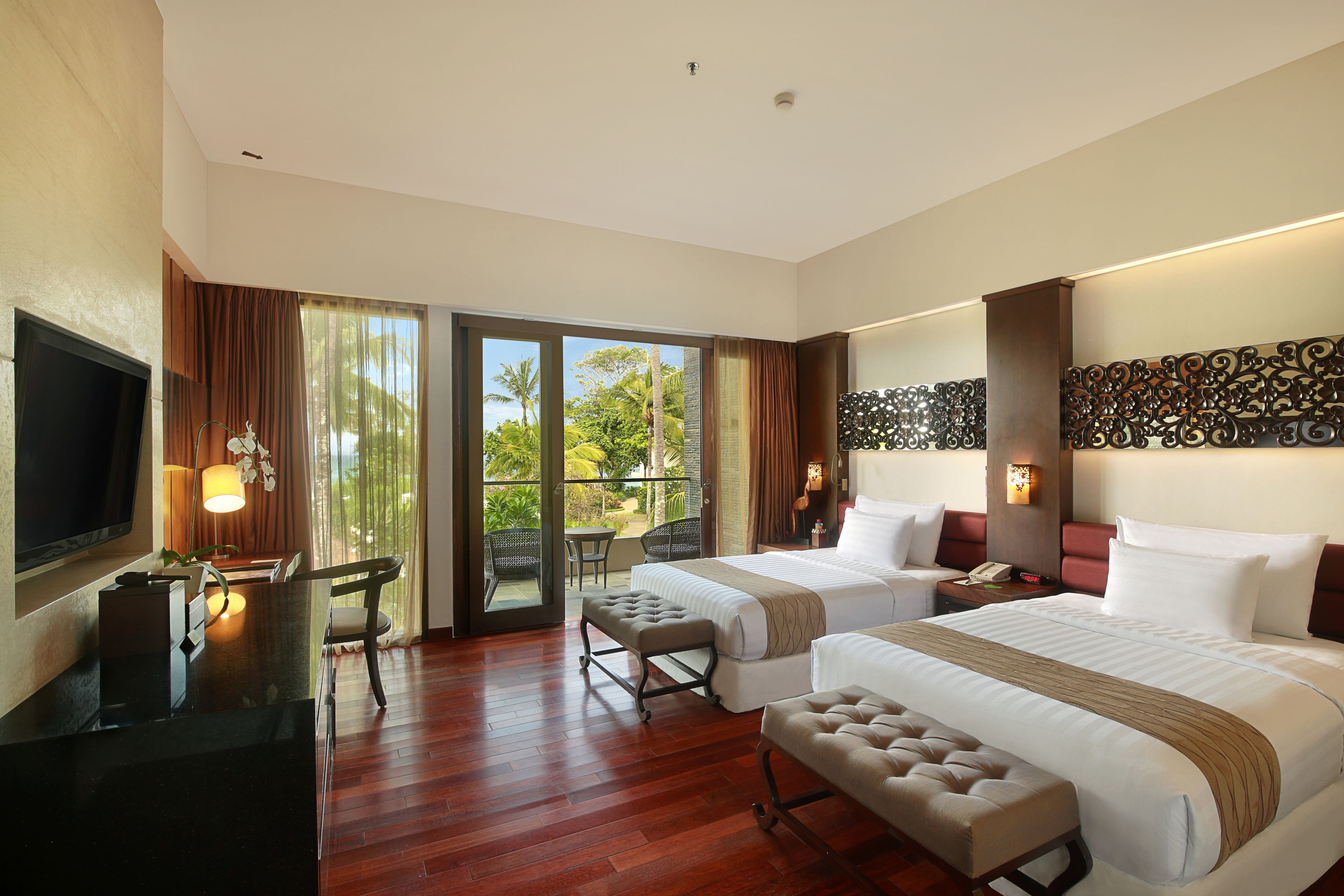 A hotel room with two beds, a TV, a desk, and a balcony with a view of greenery.