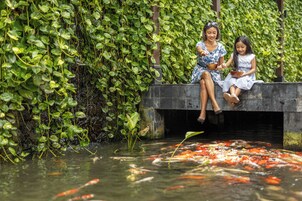 Duas pessoas sentadas num banco de pedra junto a um lago de carpas, rodeadas por vegetação exuberante.