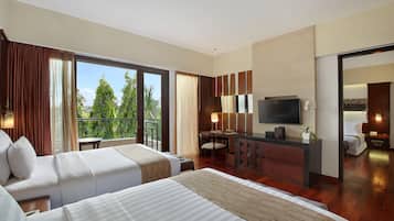 A hotel room with two beds, a desk, a TV, and a balcony with a view of trees.