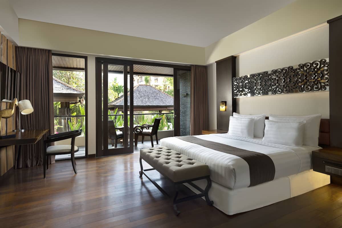 A spacious bedroom with a large bed, a desk, and a chair, a balcony with a view of the outside.