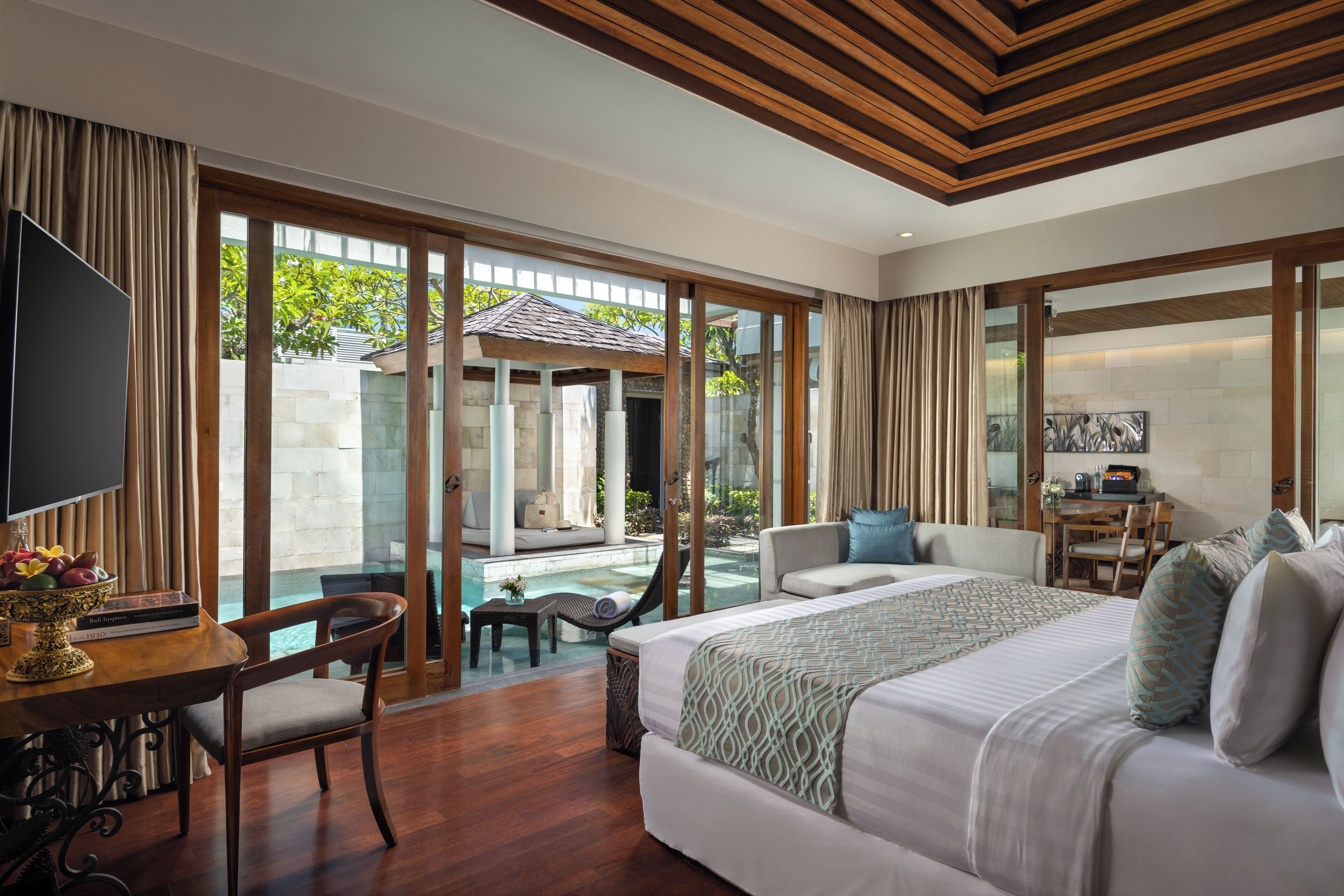 A spacious bedroom with a large bed, a TV, and a dining area with a view of a pool and outdoor seating.