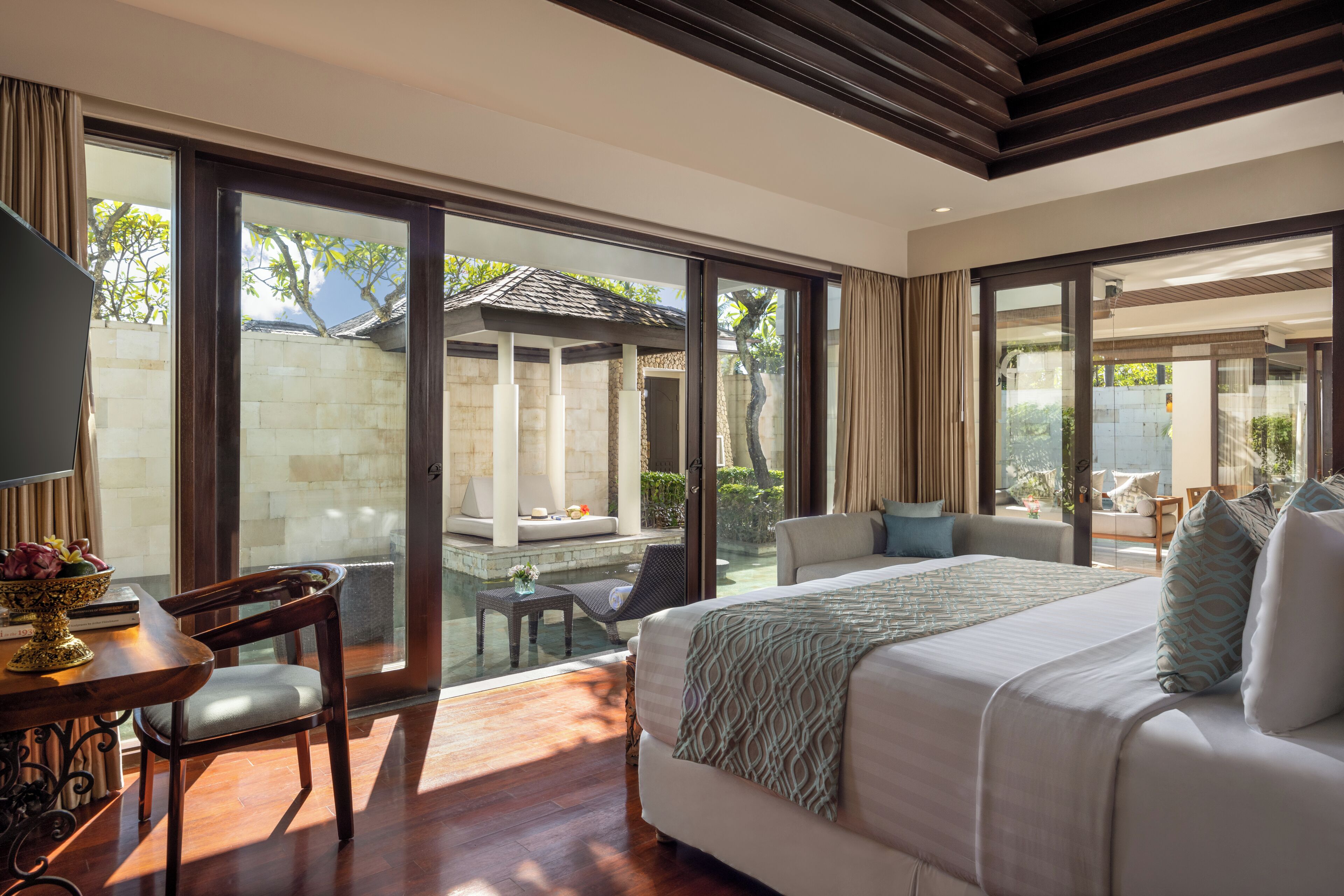 Um quarto espaçoso com uma cama grande, um espaço de estar com sofá e cadeiras, e vista para uma piscina e jardim através de grandes portas de vidro deslizantes.