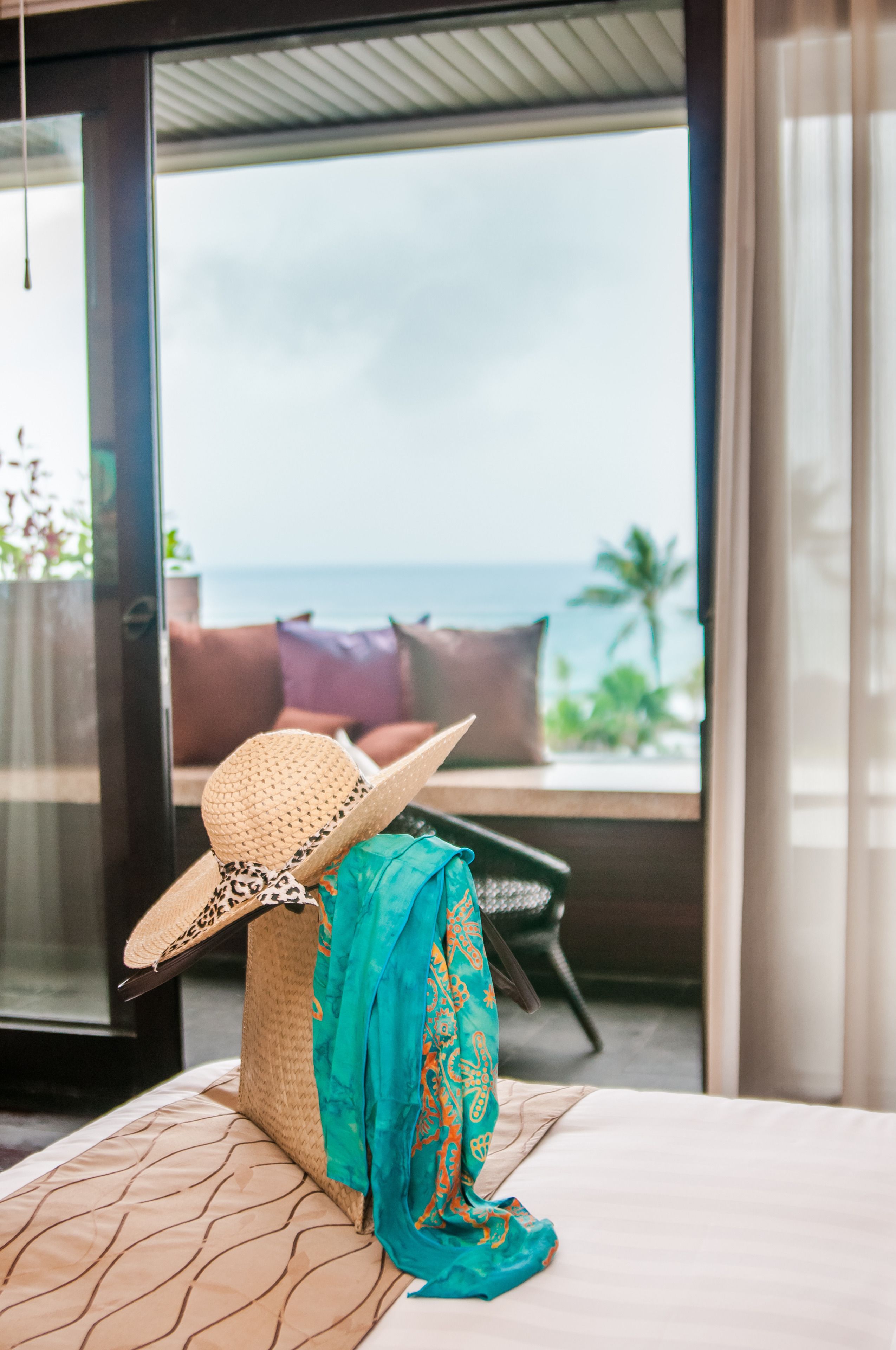 A room with a view of the ocean, a bed with a patterned bedspread, a straw hat, and a woven bag.