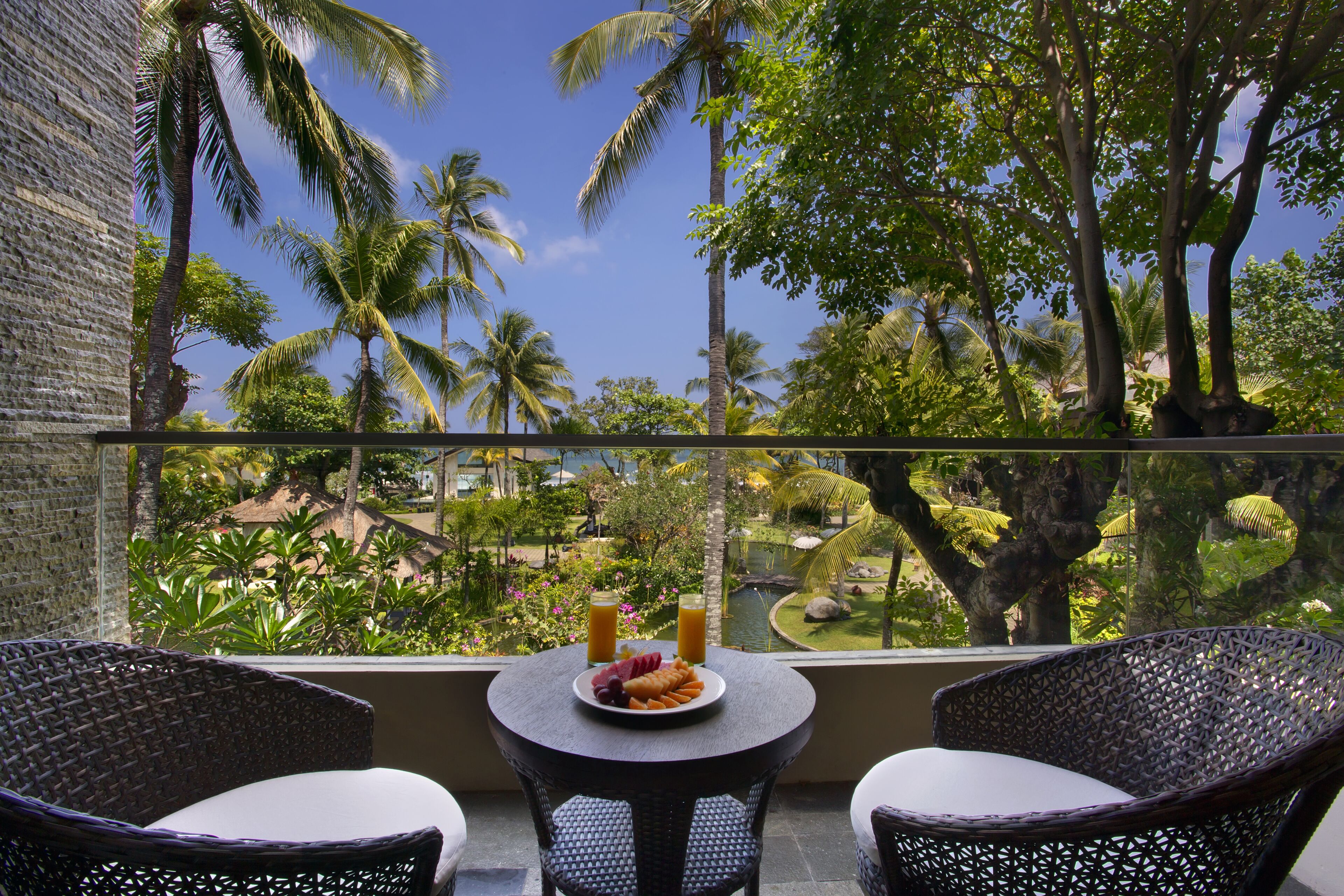 A balcony with wicker chairs, a small table with a plate of fruit and two glasses of orange juice, and a view of palm trees and a garden.