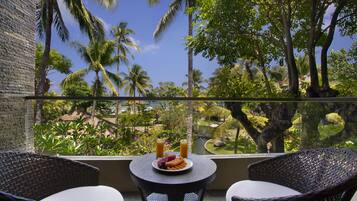 A balcony with wicker chairs, a small table with a plate of fruit and two glasses of orange juice, and a view of palm trees and a garden.