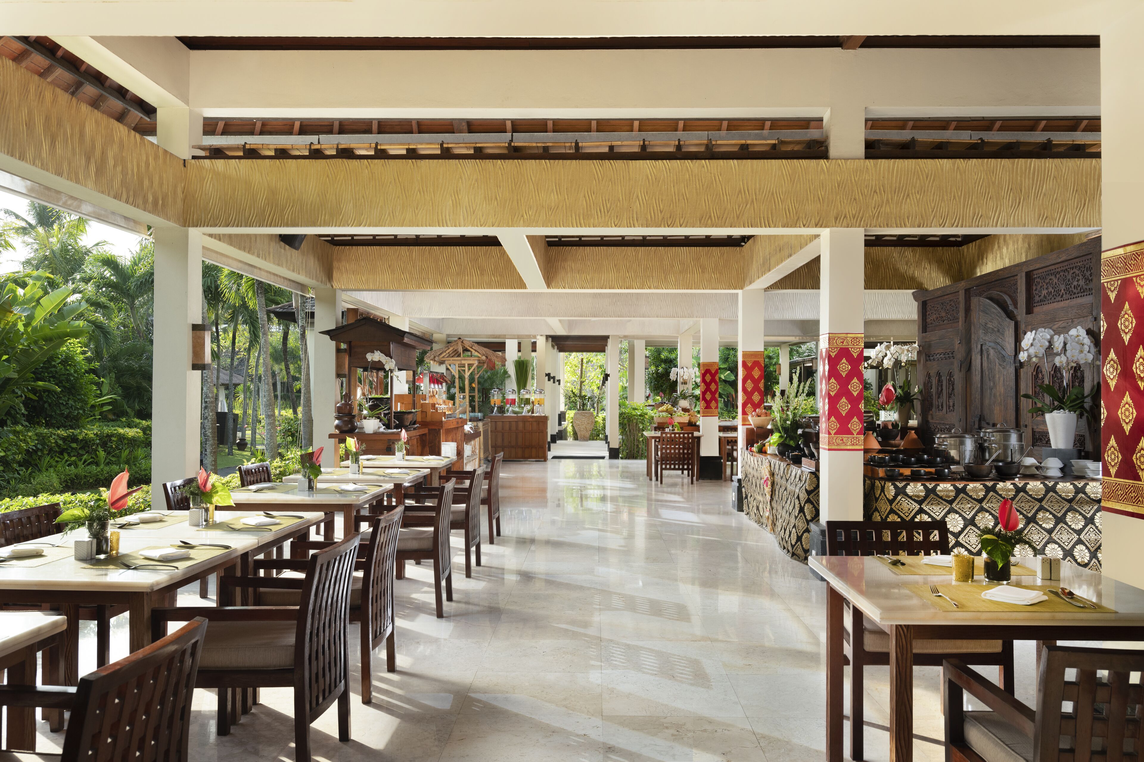 A spacious indoor dining area with high ceilings and natural light, featuring wooden tables and chairs, a central bar area, and a view of lush greenery outside.