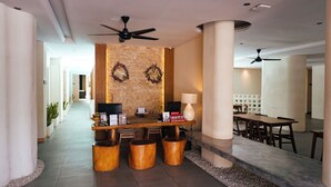 A hotel reception area with a wooden reception desk, a wall-mounted TV, a lamp, and a Christmas wreath. There are wooden stools and a seating area with tables and chairs. - The Rivavi Legian (Legian)