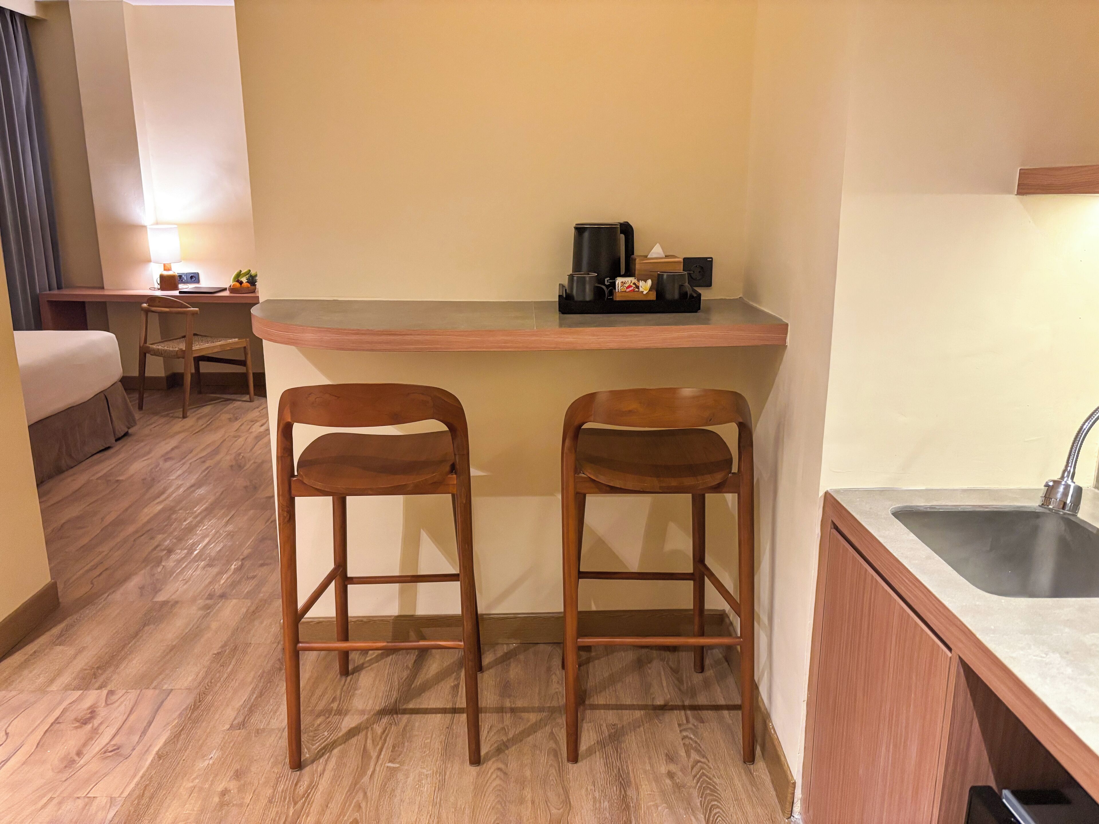 Une chambre d’hôtel compacte comprenant un coin cuisine, deux tabourets de bar en bois, un lit, un bureau avec une lampe et un lavabo.
