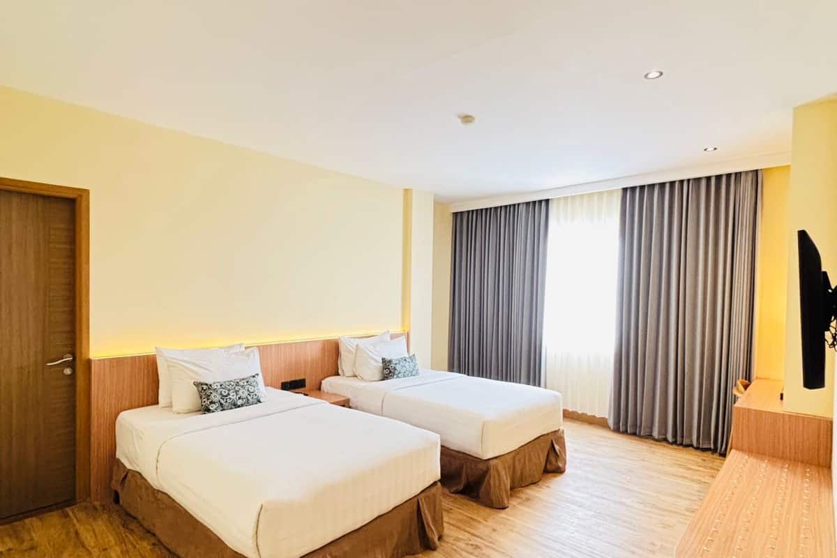 A hotel room with two beds, wooden flooring, a wooden door, a large window with curtains, and a desk with a lamp.