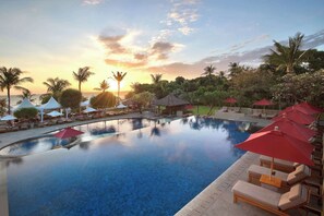 Zona de piscina con sillones de descanso y sombrillas, rodeada de palmeras y vista al atardecer.