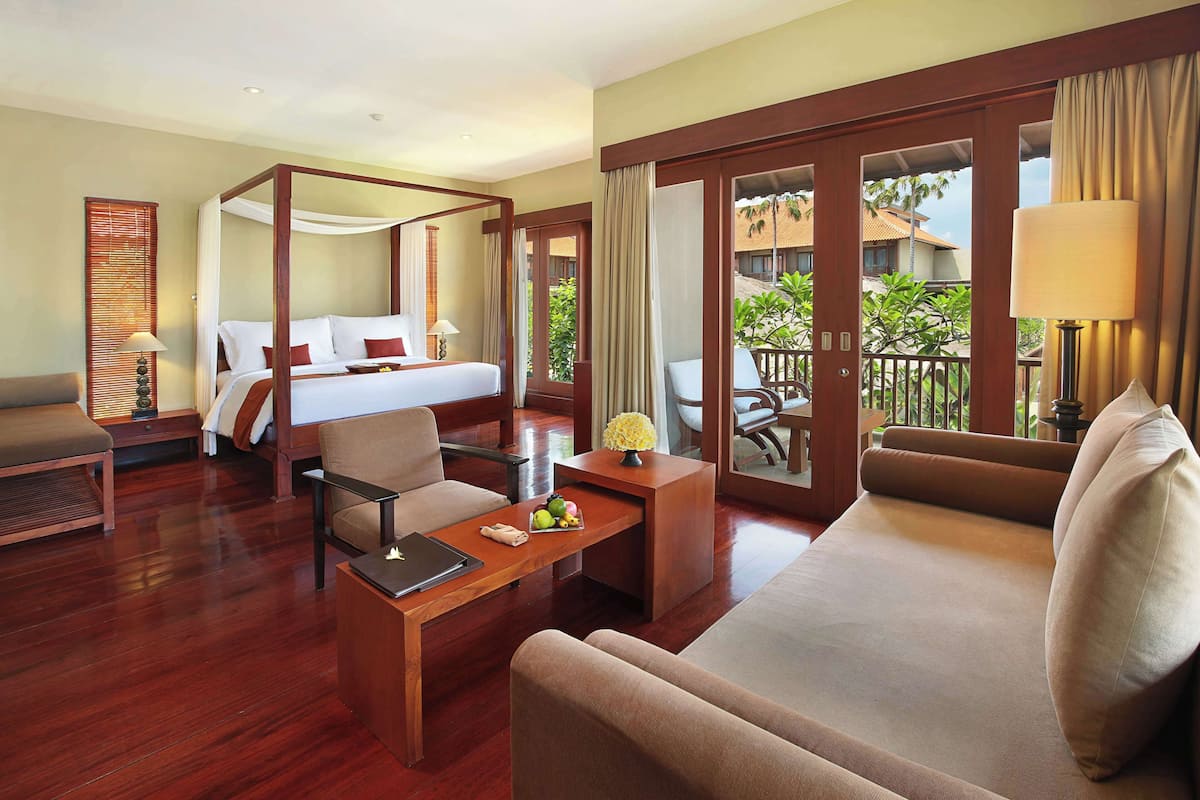 A hotel room with a four-poster bed, a sofa, a dining table, and a balcony with a view of the surrounding area.
