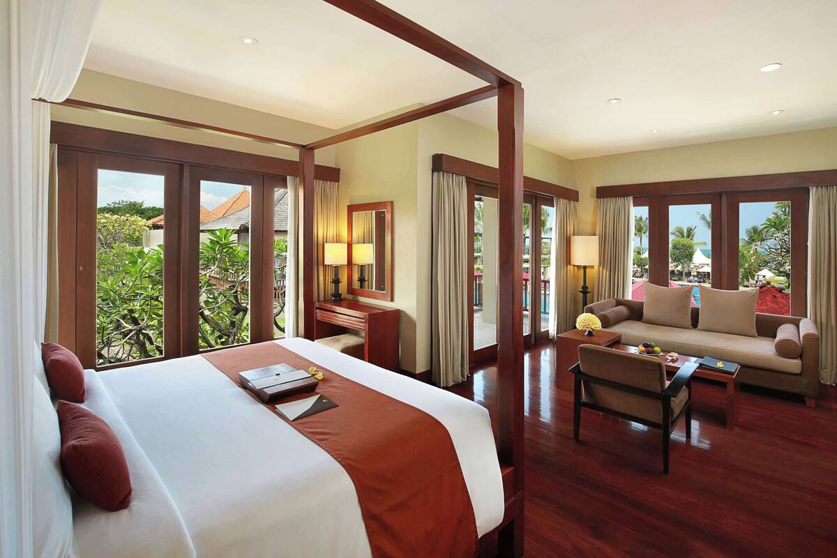 A hotel room with a large bed, a sofa, a coffee table, and a desk. There are large windows with curtains, and a view of the outdoors.