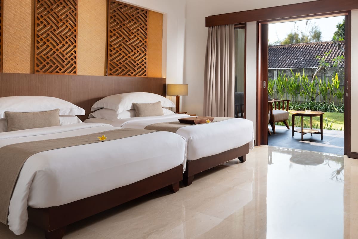 A hotel room with two beds, a wooden headboard, a balcony with a view of greenery, and a sliding glass door.