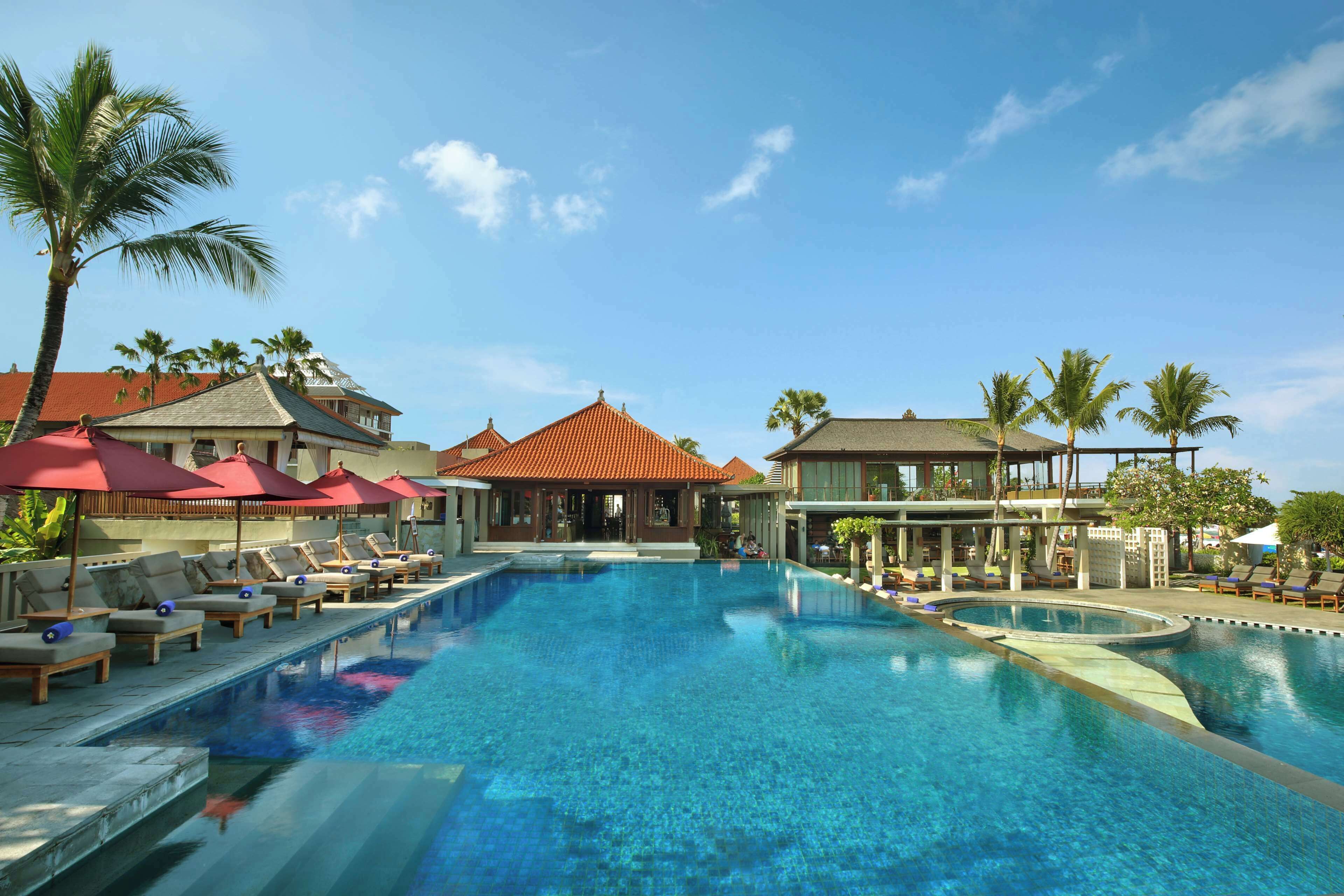 A large swimming pool with lounge chairs and umbrellas, surrounded by buildings with red roofs and palm trees.