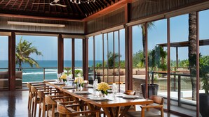 A dining area with a long wooden table set for six, large windows offering ocean views, and a ceiling fan. - Bali Niksoma Boutique Beach Resort (Legian)