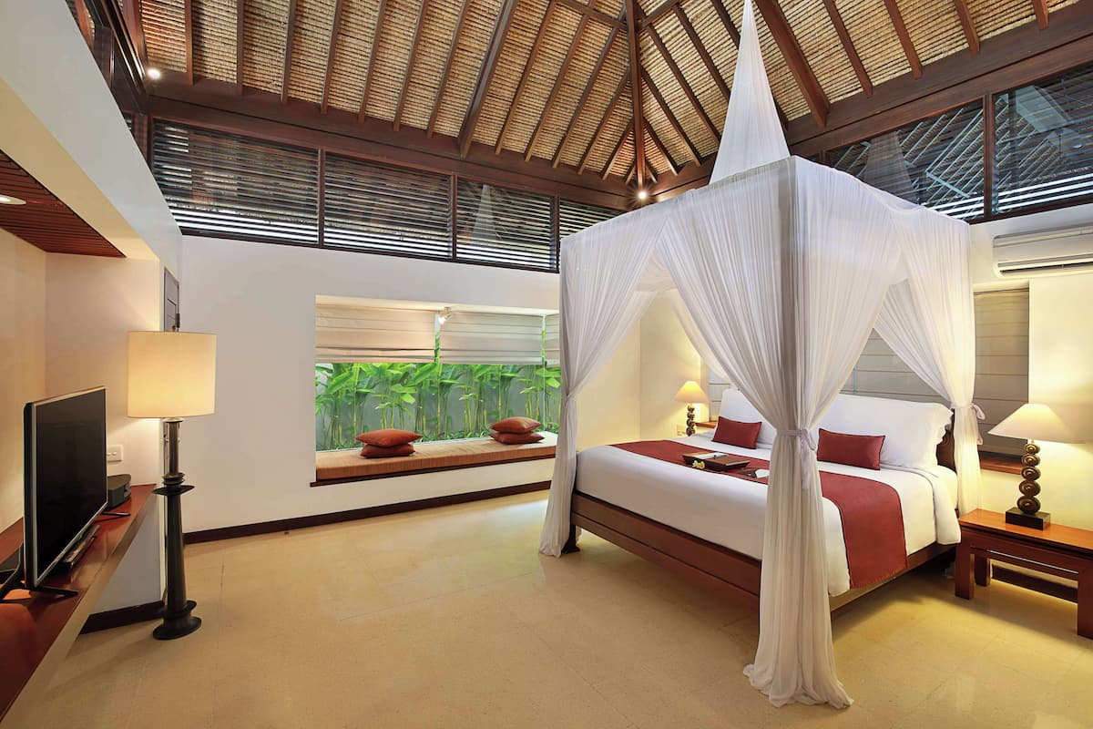 A hotel room with a bed, a TV, a bedside table, a lamp, and a window with a view of greenery.