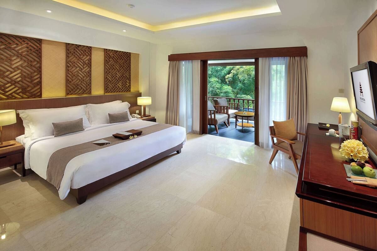 A hotel room with a large bed, a desk with a TV, and a balcony with a view of trees.