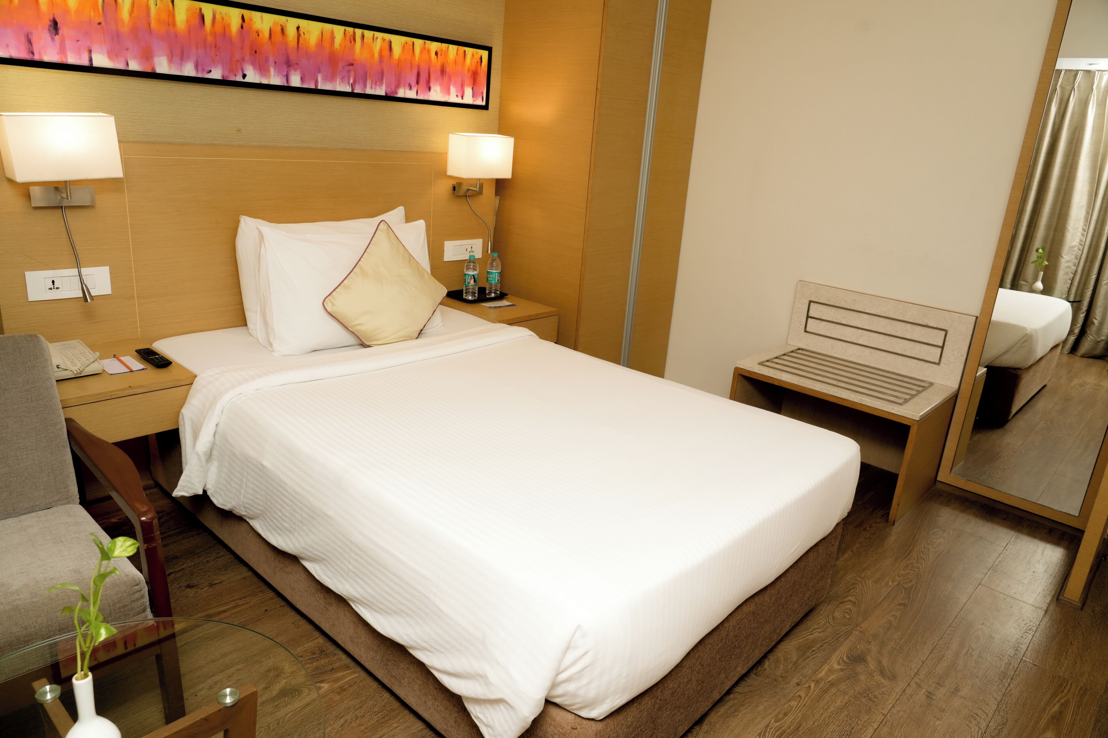 A hotel room with a bed, bedside tables, a desk, a chair, a glass coffee table with a plant, and a large mirror.