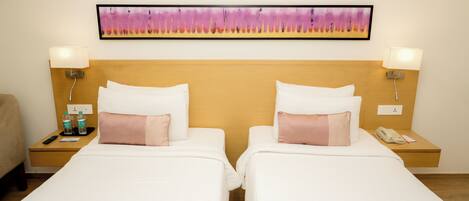 Habitación de hotel con dos camas, cada una con ropa blanca y almohadas rosas, un cabecero de madera, un cuadro sobre las camas y lámparas de noche.