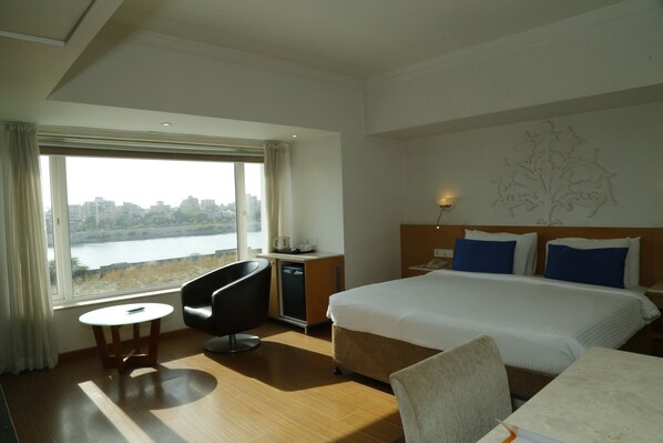 Habitación de hotel con una cama grande, un escritorio, una silla y vistas a un paisaje urbano a través de un ventanal.