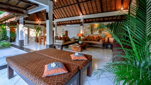 A spacious living area with a large bed, sofas, and a coffee table. There are multiple plants and a large window providing natural light. - The Maya Seminyak Villas (Seminyak)