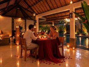 A couple dining at a table with candles and rose petals on a tiled floor, overlooking a pool. - The Maya Seminyak Villas (Seminyak)