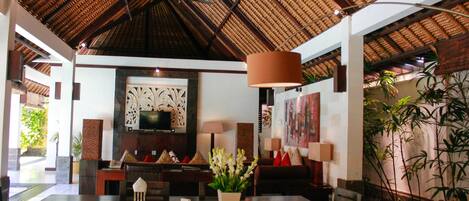A spacious room with a high ceiling and a thatched roof, featuring a dining area with a table and chairs, a sofa, and a television.