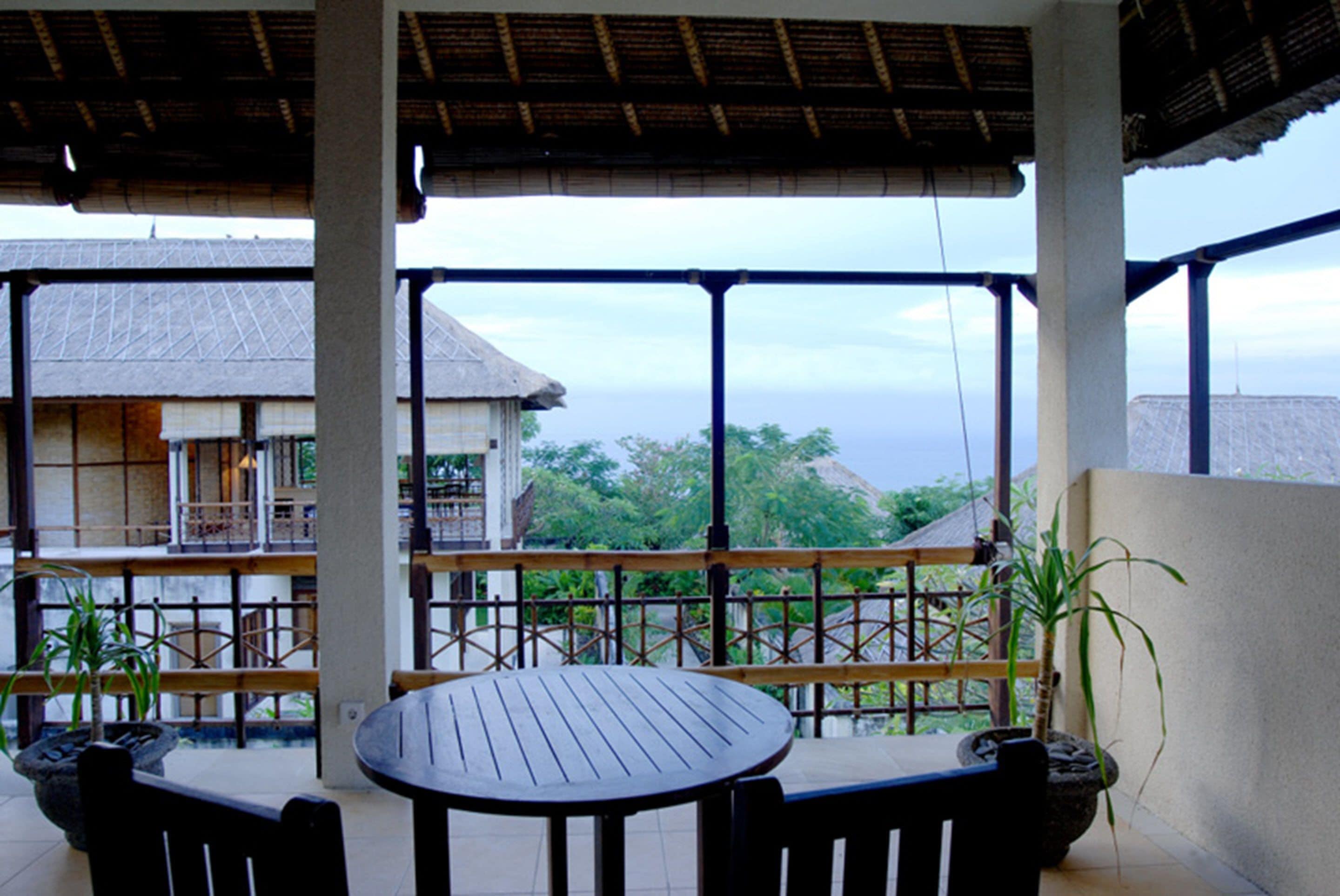 A covered outdoor seating area with a table and chairs, overlooking a building and a view of the ocean.