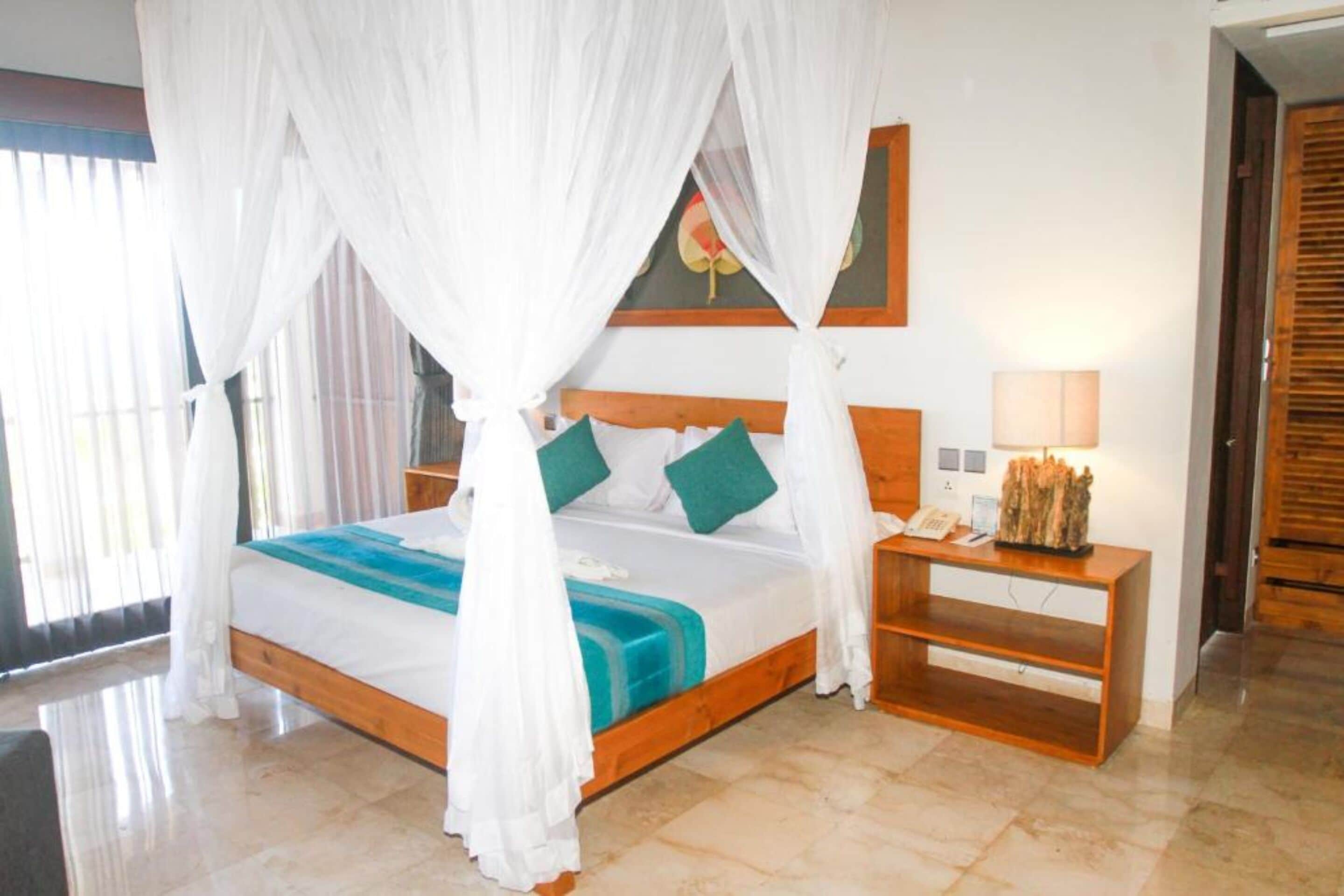 A bedroom with a wooden bed, white canopy, teal and white bedding, a nightstand with a lamp, and a painting on the wall.