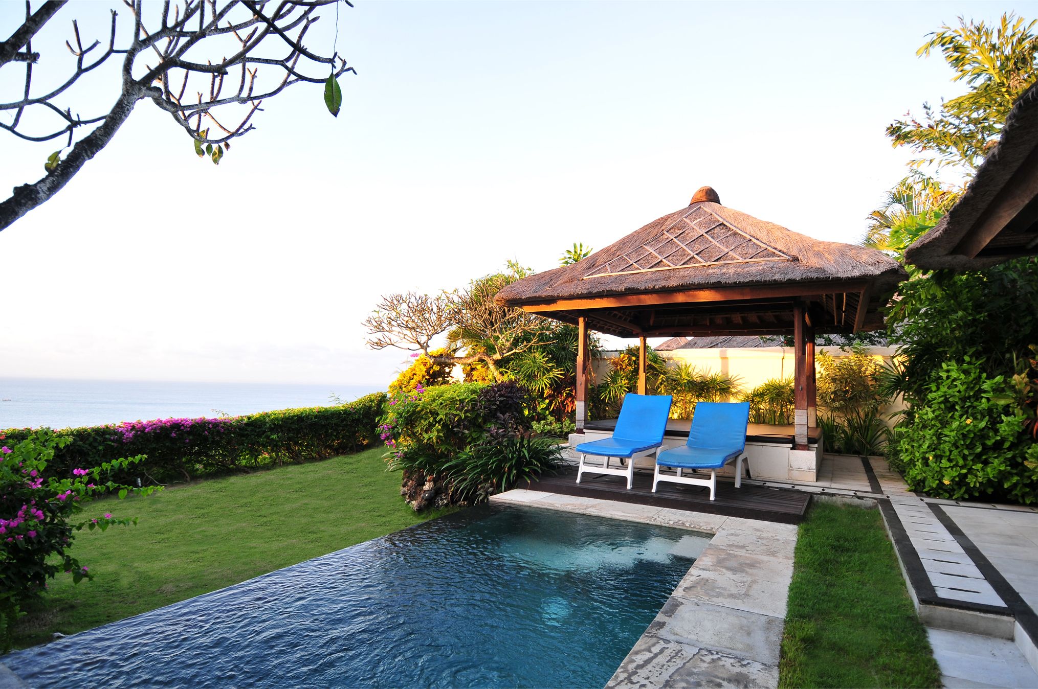 A pool area with lounge chairs, a thatched gazebo, and a view of the ocean.