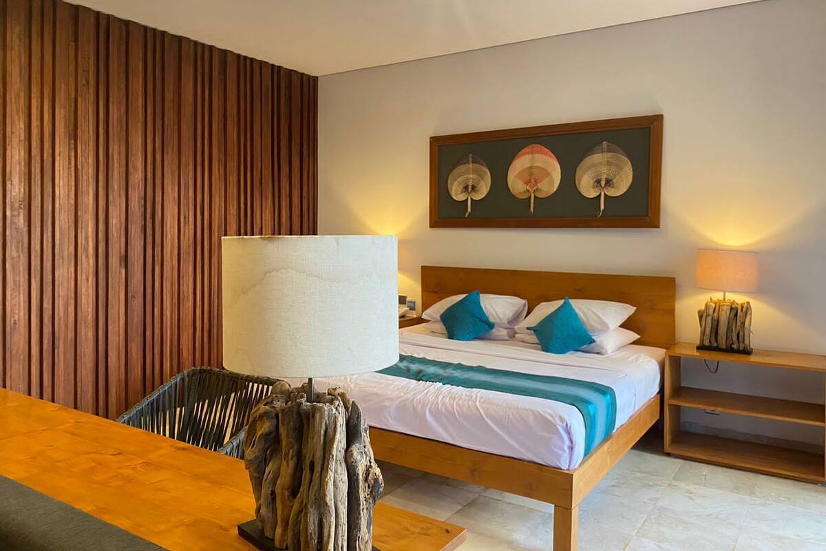A hotel room with a wooden headboard, a bed with white and teal bedding, a wooden side table with a lamp, a wooden chair, and a wall-mounted fan.