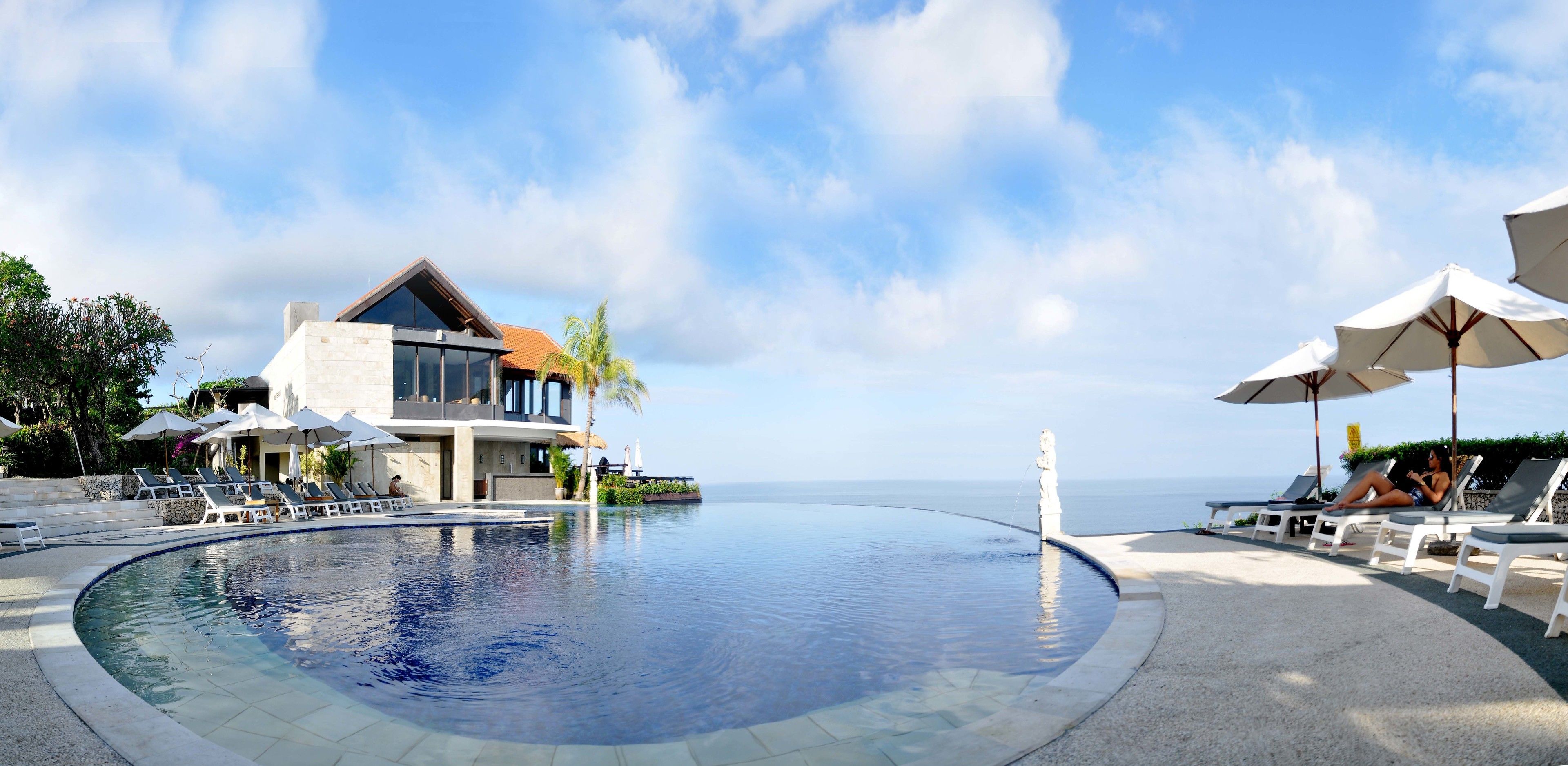 A poolside area with lounge chairs and umbrellas, a modern building with a glass facade, and a view of the sea.