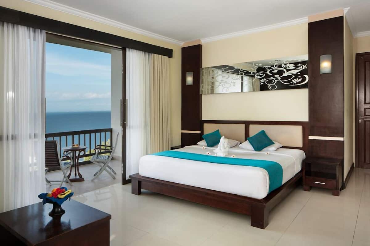 A hotel room with a large bed, a balcony with a view of the sea, a small table with a vase, and a mirror on the wall.