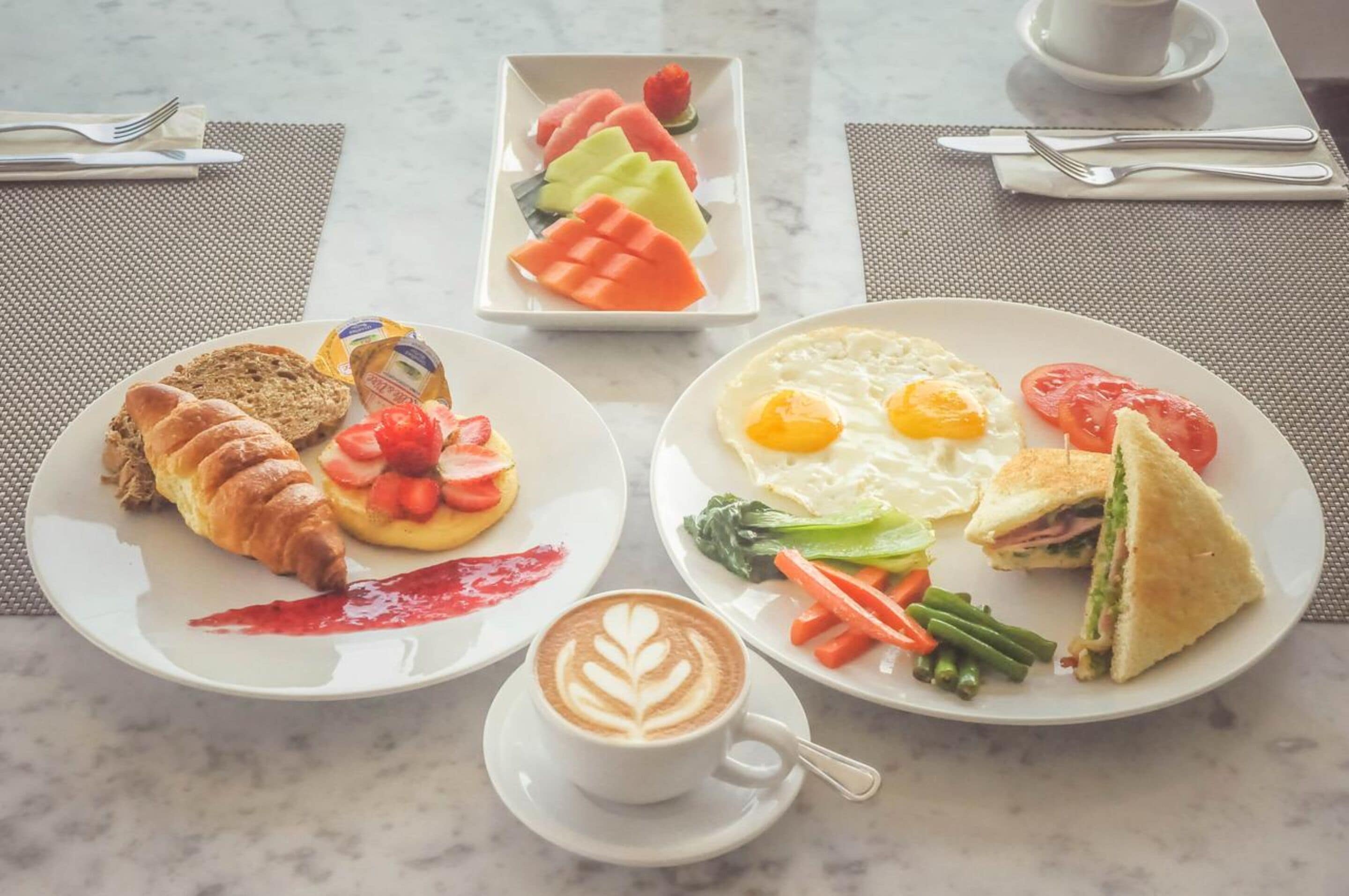 Een ontbijtsetting met croissants, aardbeien, een latte, een bord met eieren, tomaten en een sandwich.
