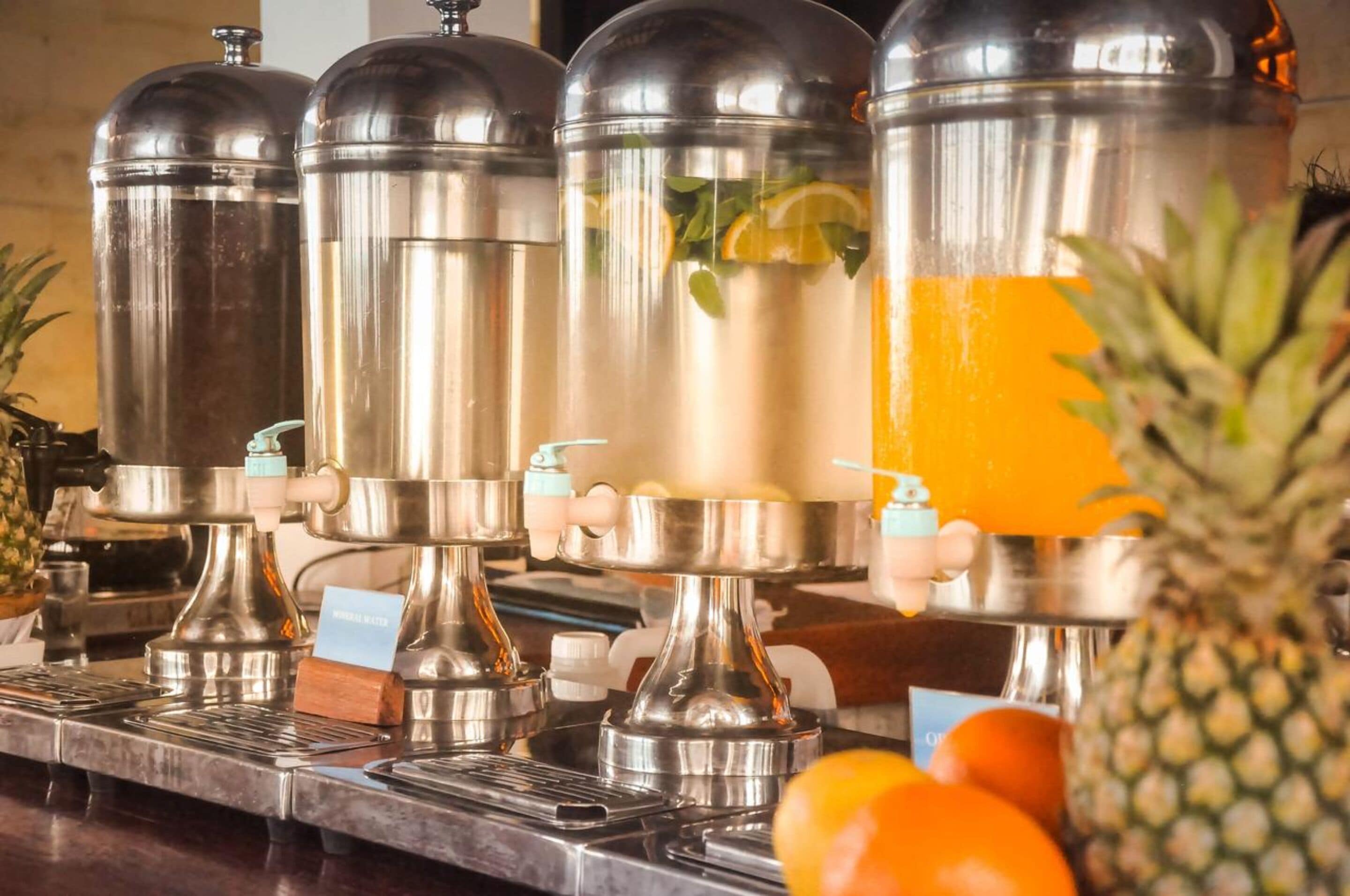 A row of stainless steel beverage dispensers with labels, a pineapple, and oranges on a counter.