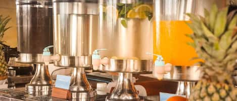 Una hilera de dispensadores de bebidas de acero inoxidable con etiquetas, una piña y naranjas sobre una encimera.