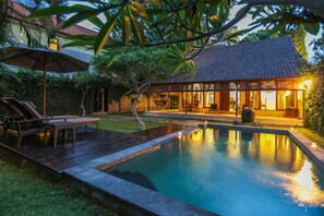 Une piscine avec une terrasse en bois et des chaises longues, entourée de verdure et bordée d’une maison dont le toit est recouvert de tuiles.