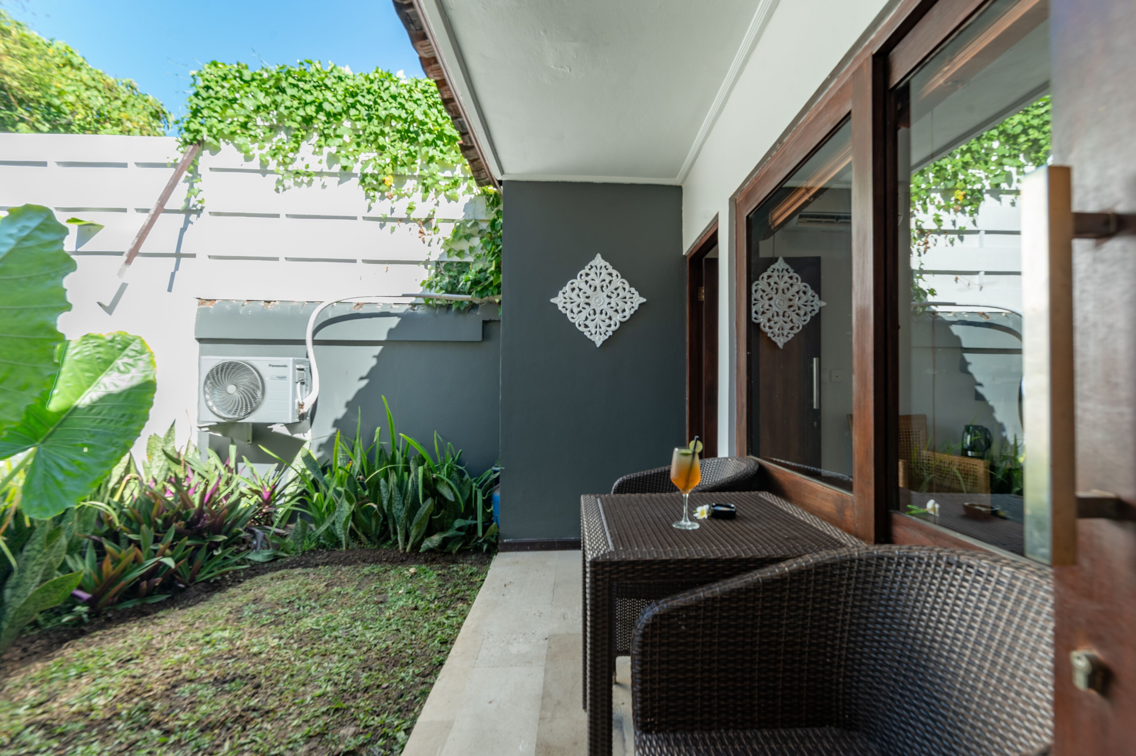Un patio con mesa y sillas de mimbre, un adorno de pared y un jardín con diversas plantas.