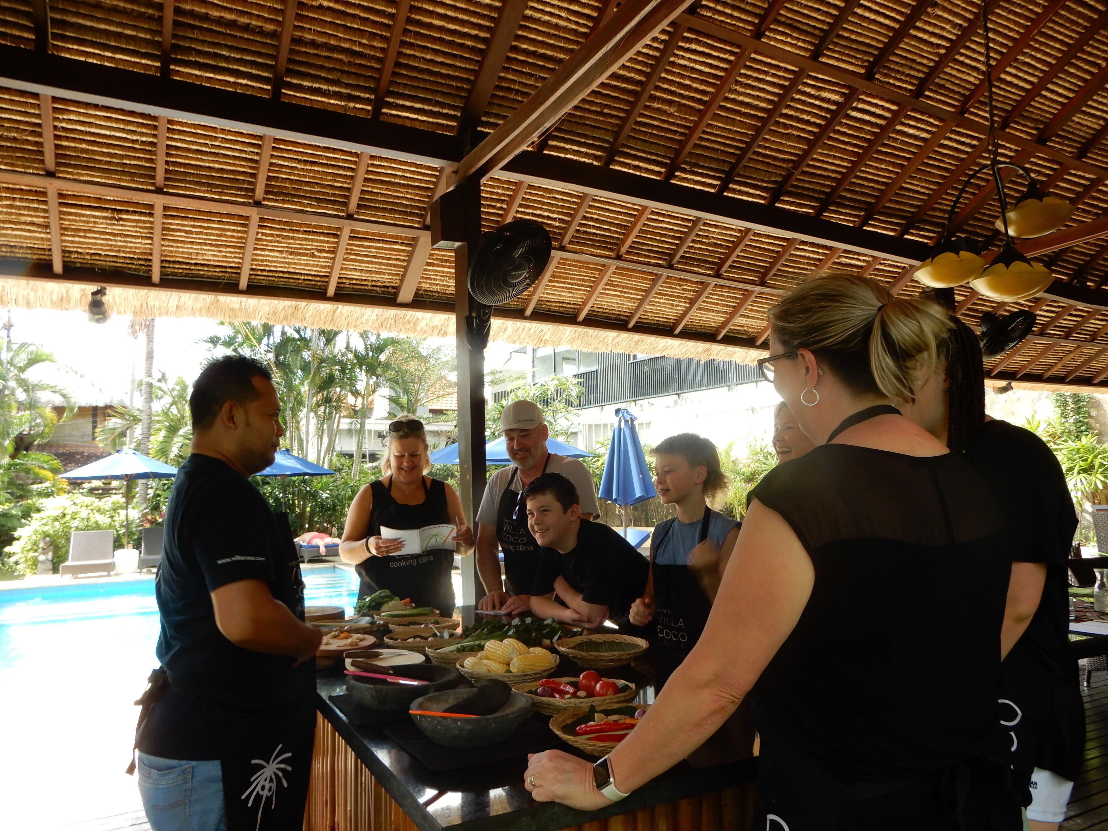 A group of people at a food preparation station with a thatched roof, surrounded by outdoor seating and a pool.