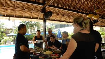 A group of people at a food preparation station with a thatched roof, surrounded by outdoor seating and a pool.