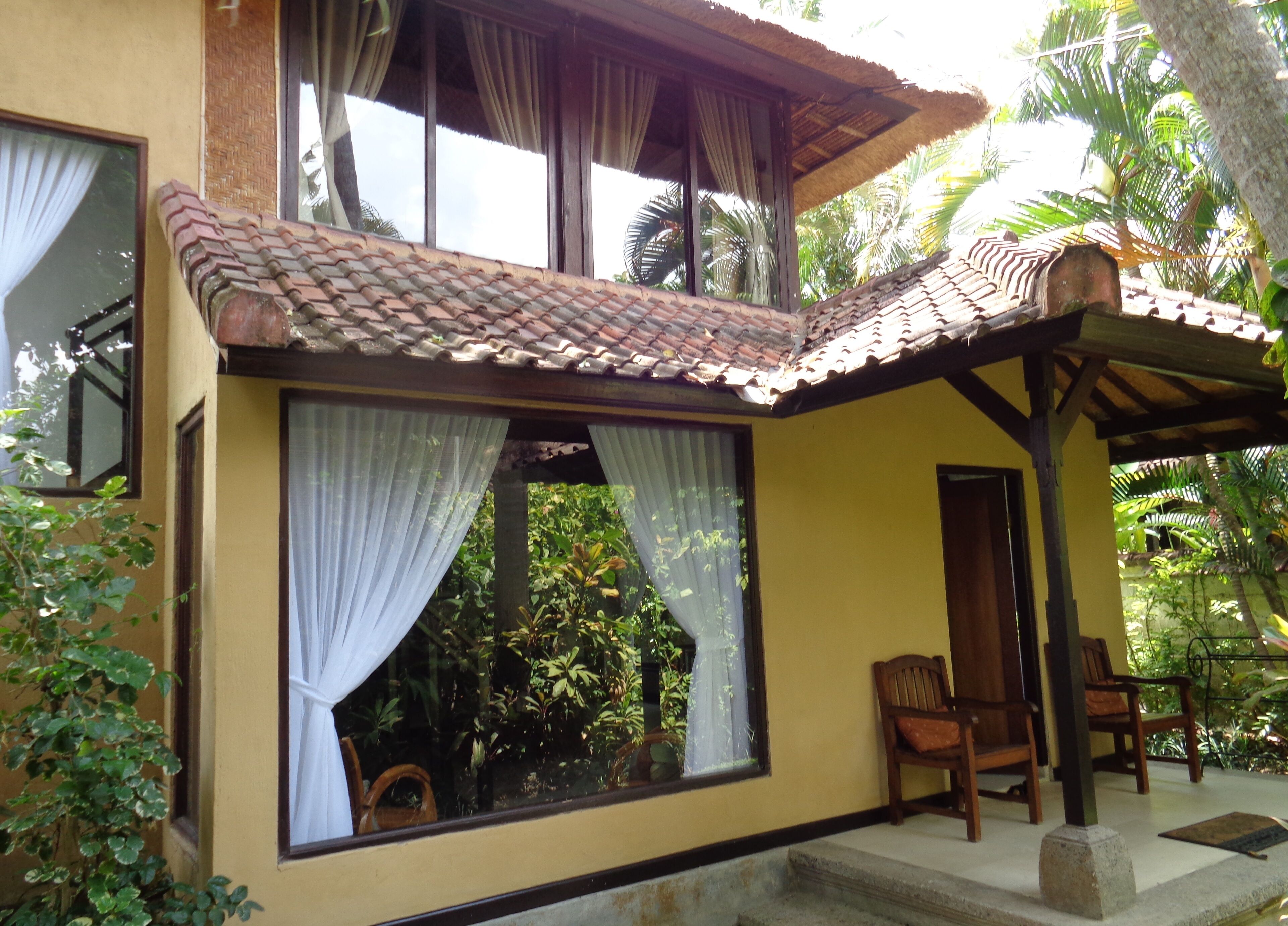 A house with a tiled roof, large windows with white curtains, and a porch with wooden chairs.