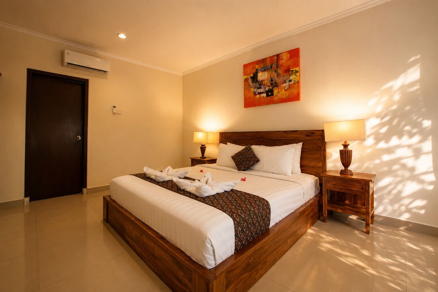 A hotel room with a large bed, two bedside tables, an air conditioning unit, and a painting on the wall.