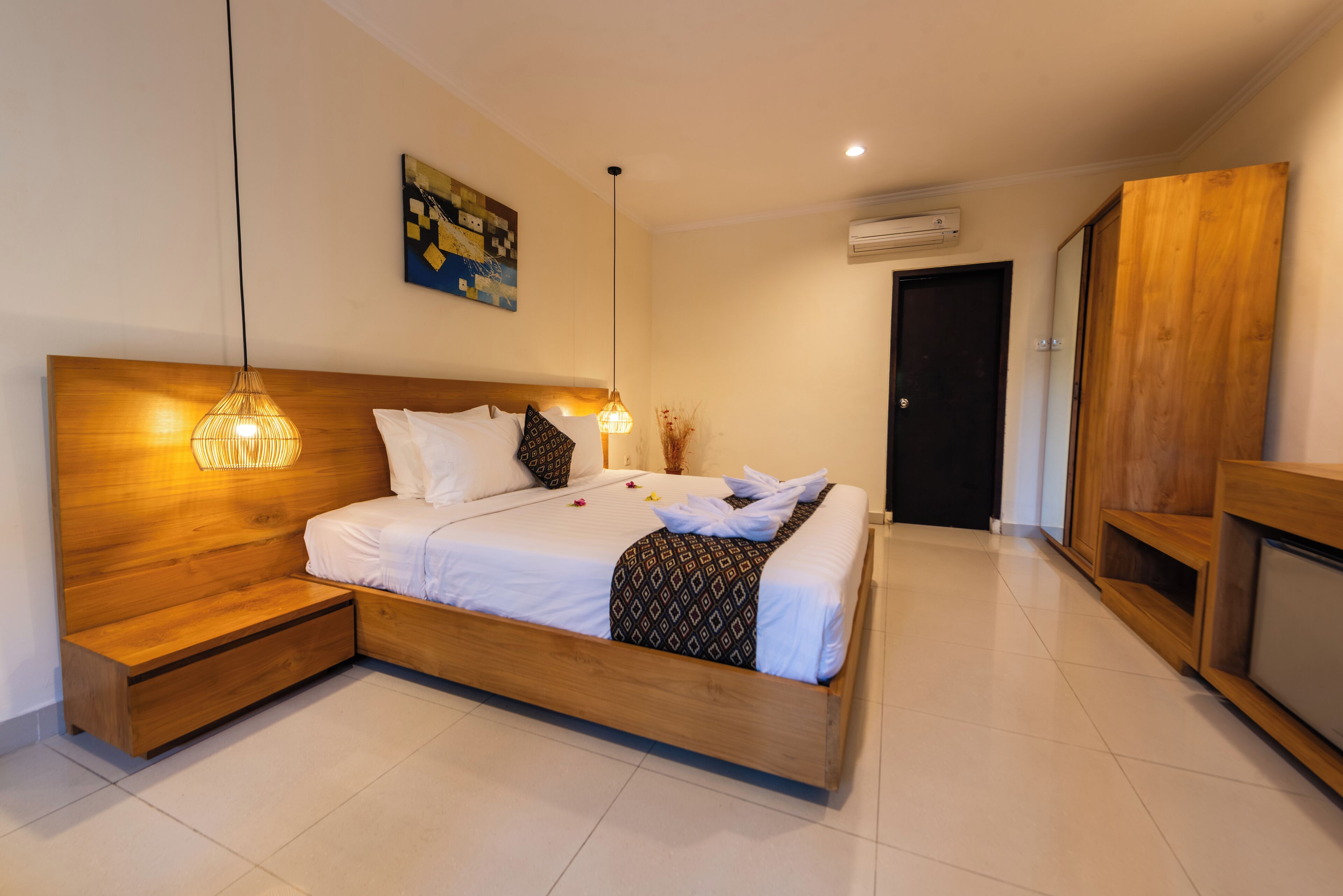 A modern bedroom with a wooden headboard, a bed with white linens, a bedside table, a hanging light fixture, and a painting on the wall.